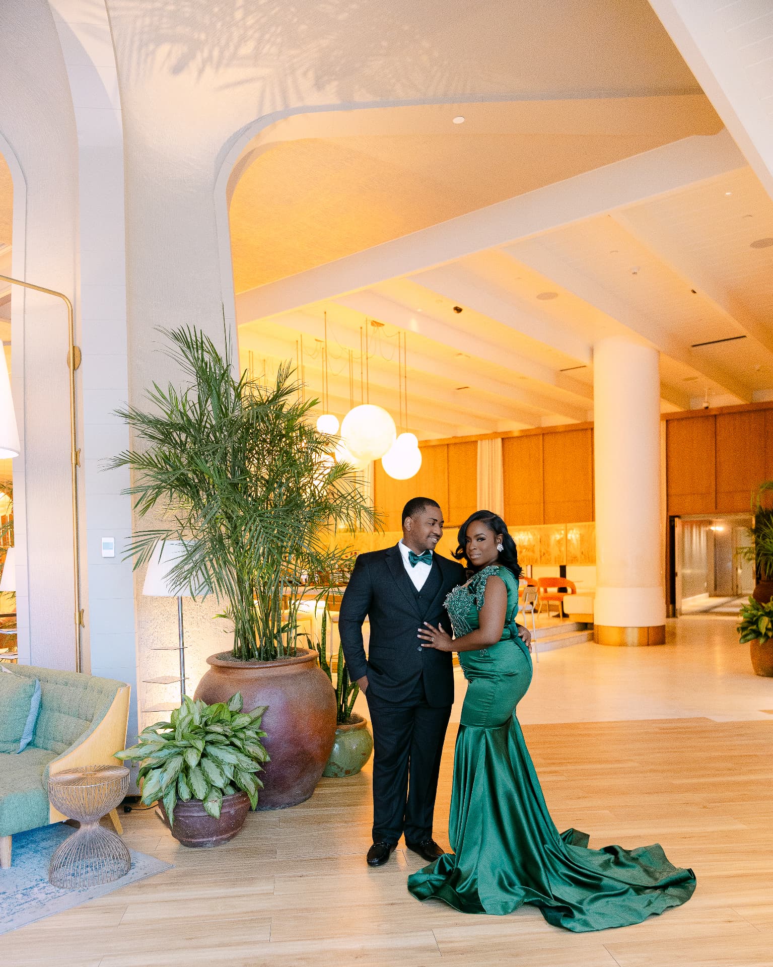 Couple in a sunlit modern hotel lobby, bride in emerald satin