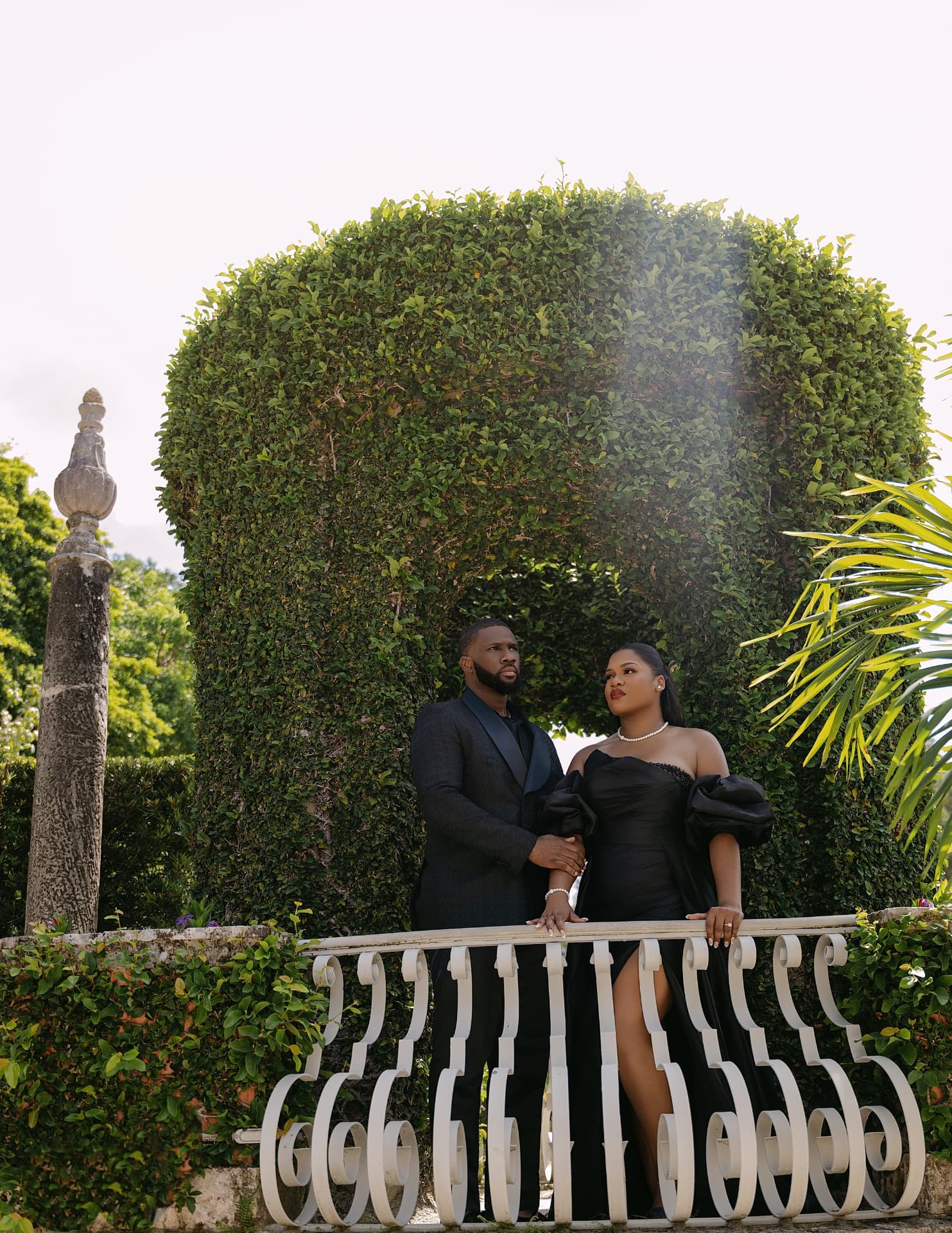 Couple in black formalwear on a scrolled balcony beneath a topiary arch