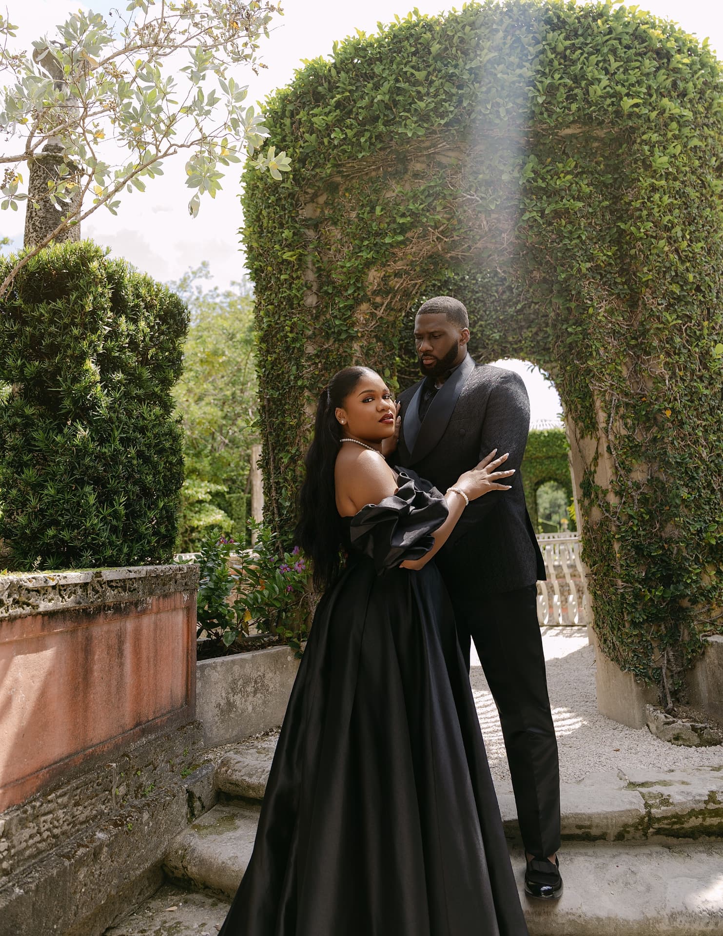 Couple in black gown and tuxedo on stone steps beneath an ivy arch