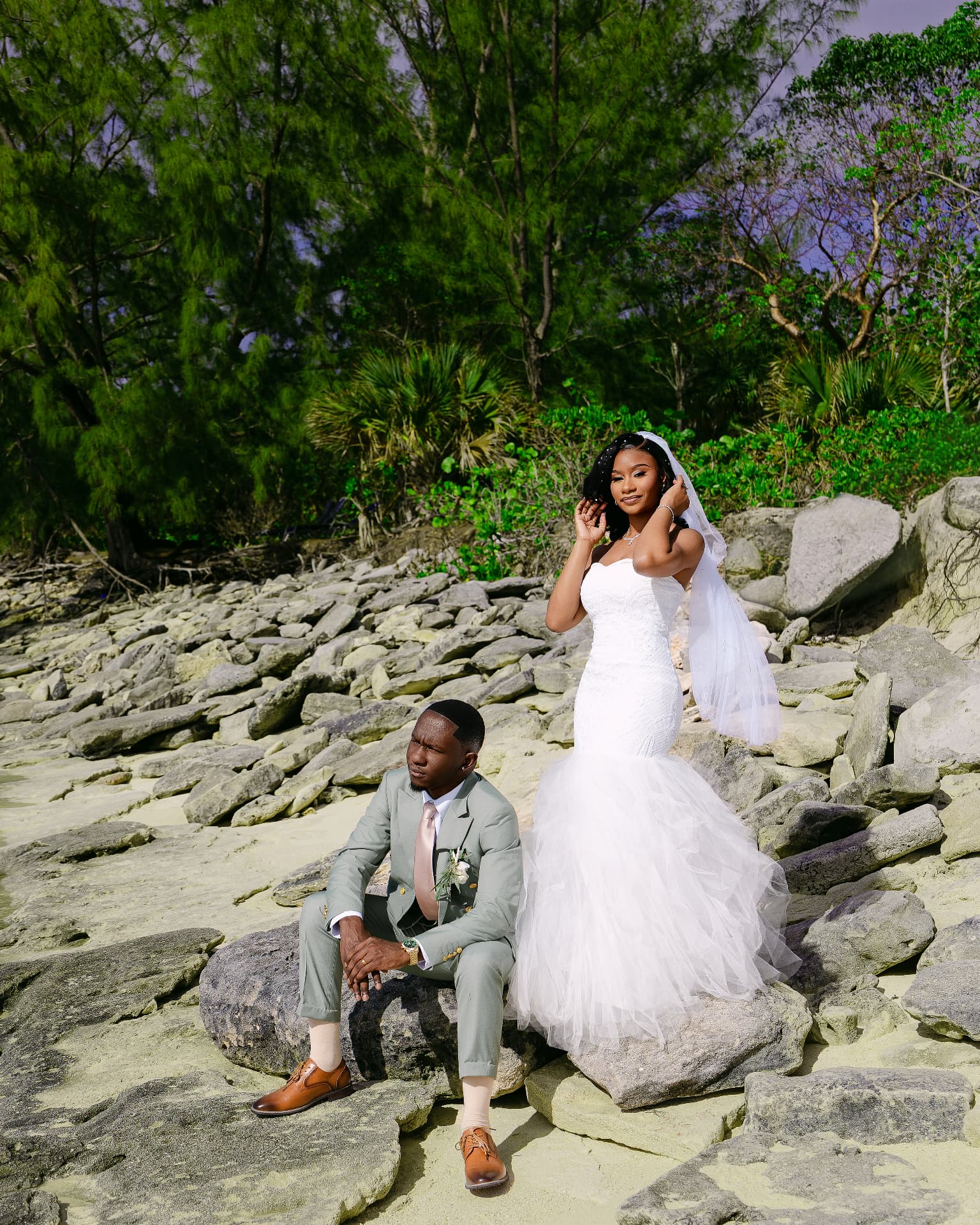 Bride with a flowing veil beside her groom on a rocky beach