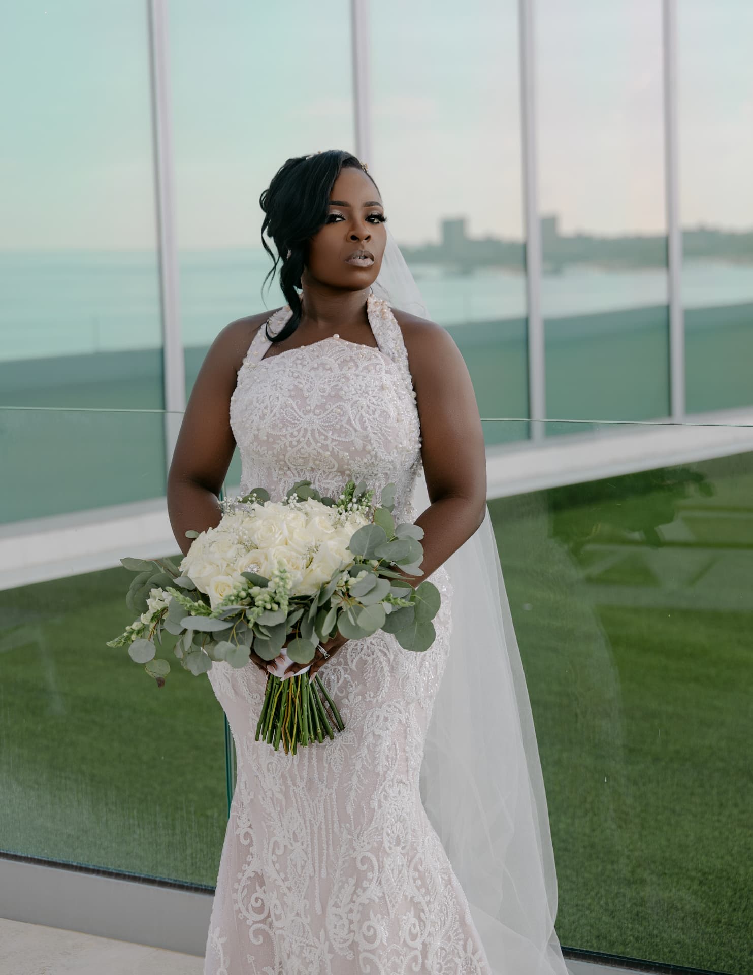 Bride in an elegant white lace gown holding a bouquet on a modern glass terrace overlooking water